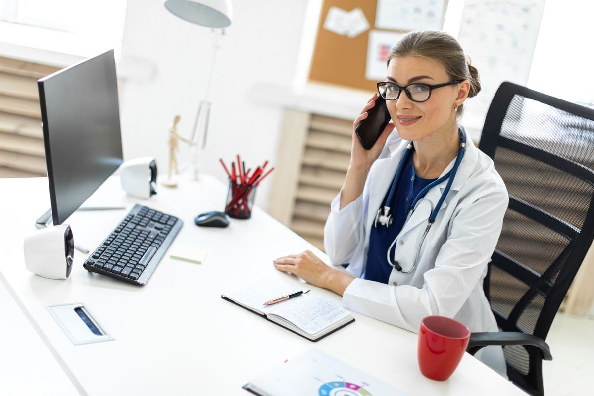 A young girl in a white robe is sitting at a table in the office and is talking on the phone. A stethoscope hangs around her neck.